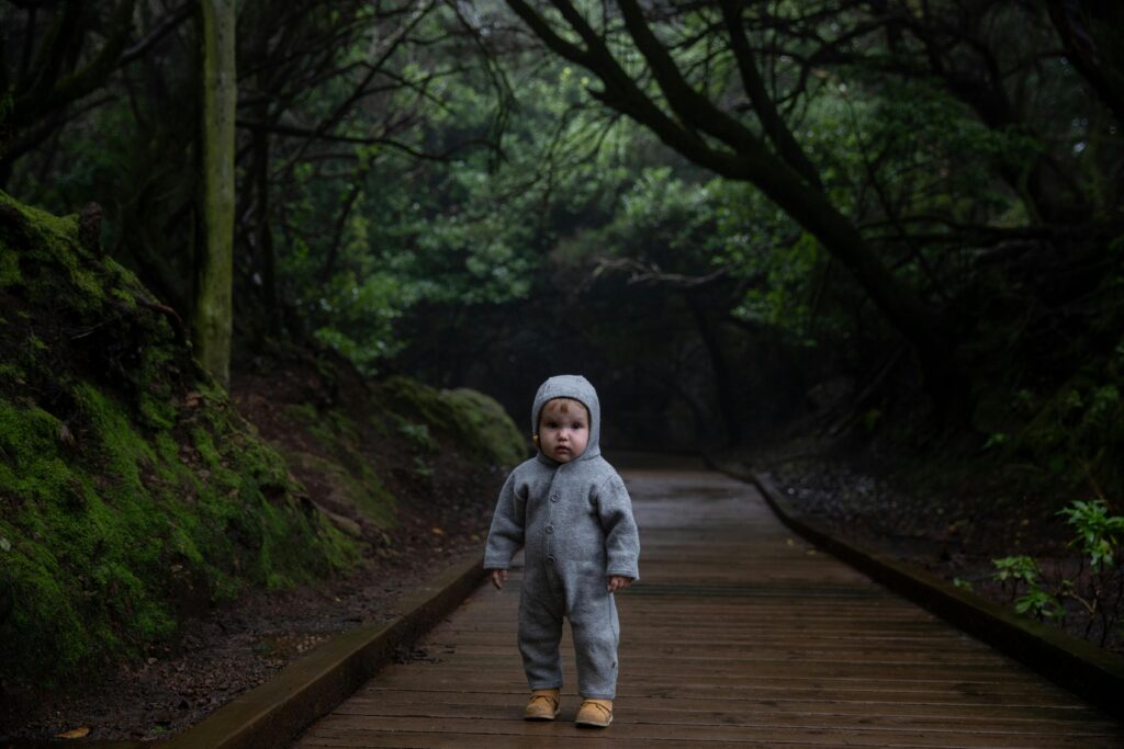 A baby in a gray outfit standing alone on a wooden pathway in a lush forest setting.